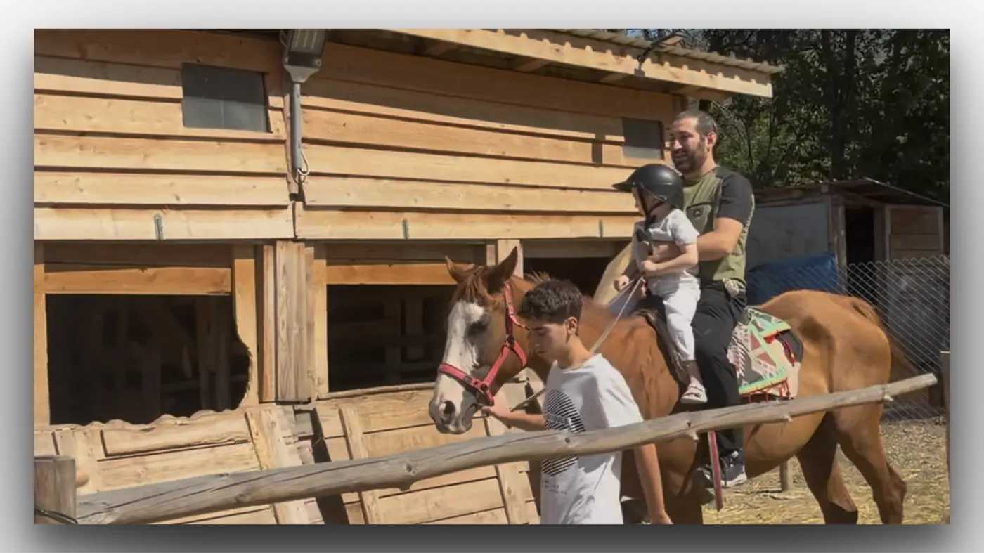 A child on horseback in front of a timber stable with an adult standing alongside