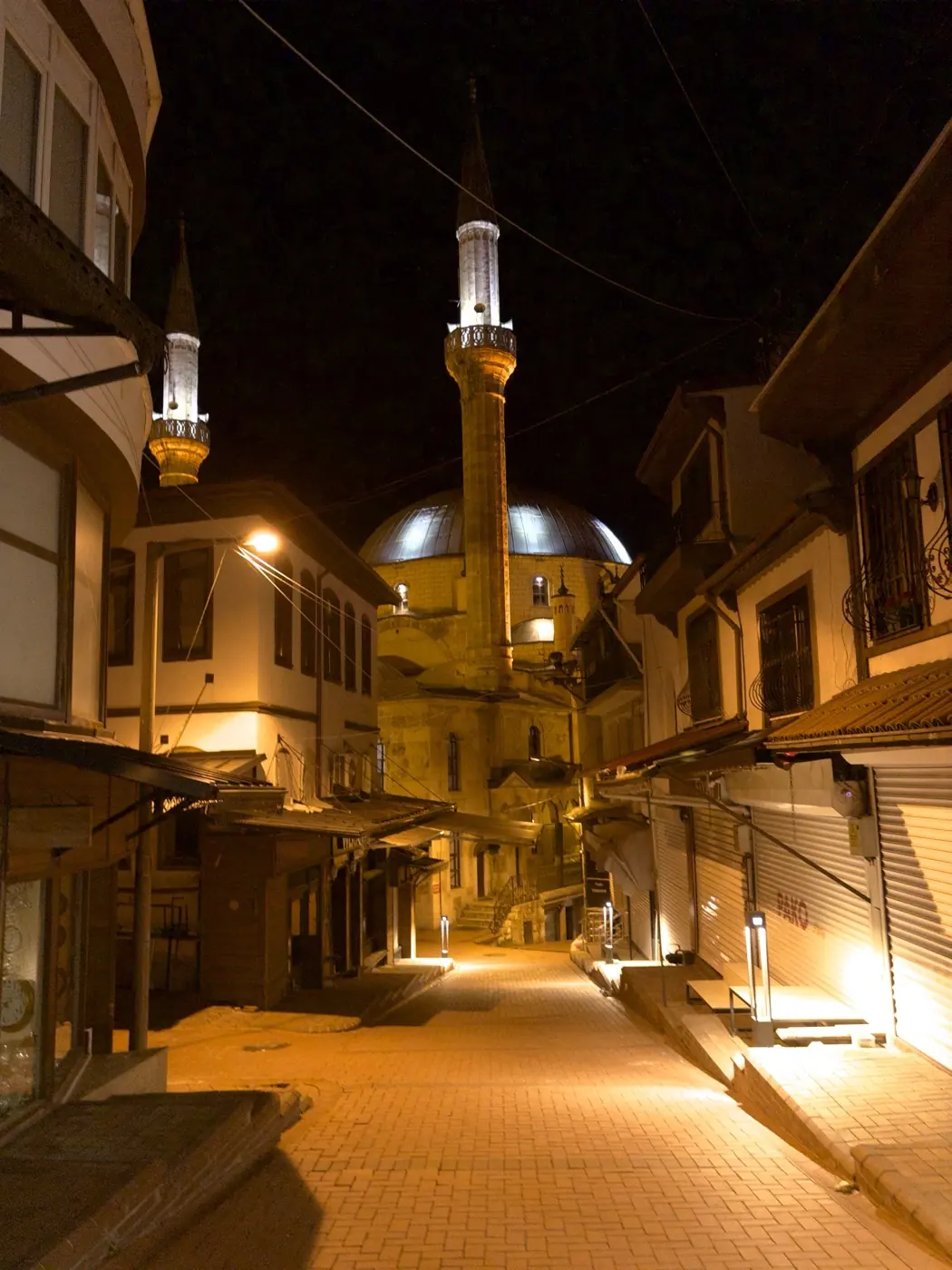 A bazaar street lit at night, shuttered shops on both sides and the two-minaret domed Bayezid Mosque at the end of it